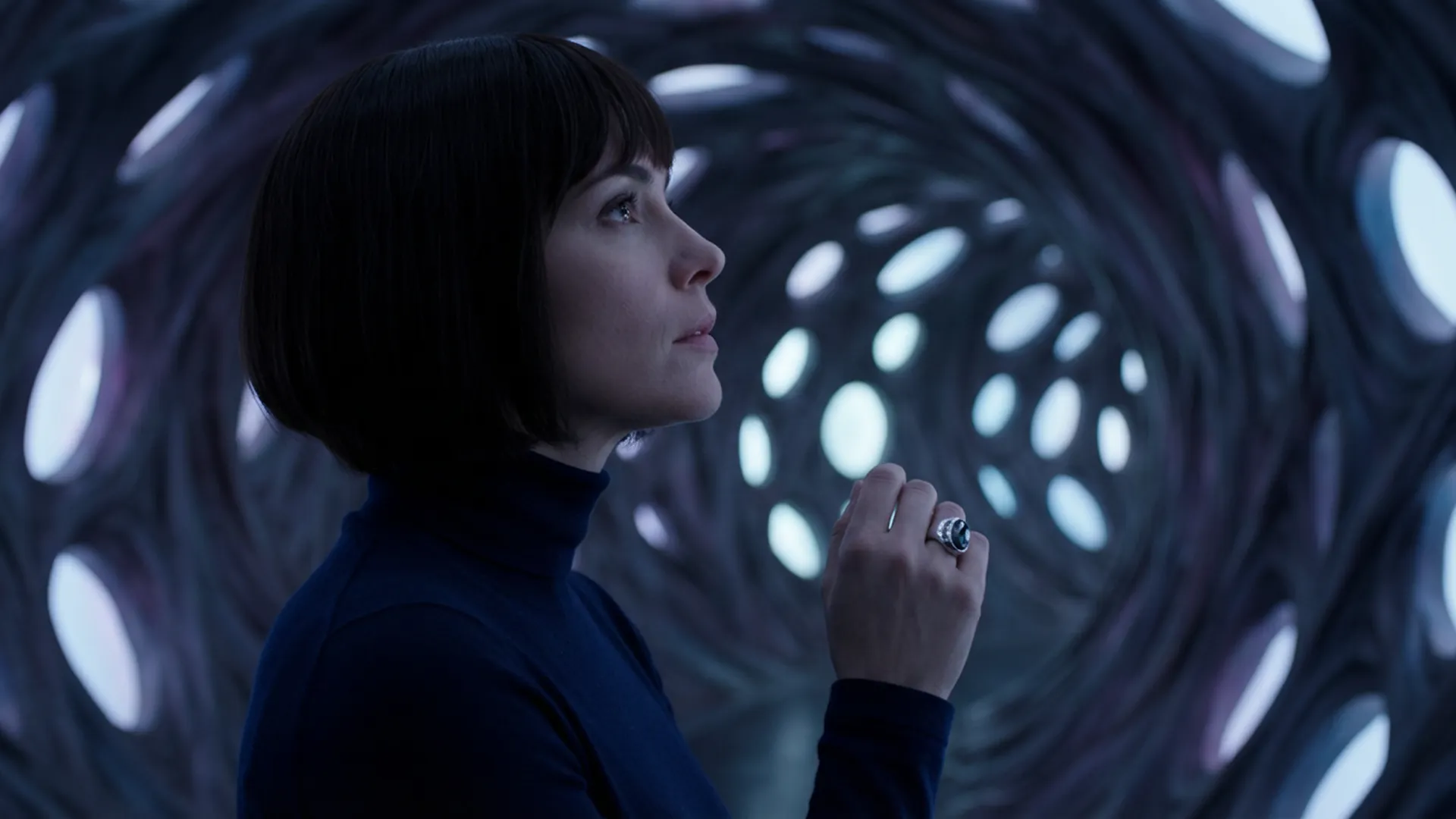 This cinematic still captures a striking portrait of a woman, rendered with a dramatic, almost theatrical flair. The camera angle is a medium shot, framing her from the waist up, with her profile turned slightly upwards and to the right, as if in contemplation or awe. Her dark, sleek bob haircut frames a delicate face with a prominent nose and full lips, her gaze directed towards an unseen wonder above and beyond the frame. She wears a simple, dark turtleneck, which allows her face and the intricate background to take center stage. Her right hand is raised, fingers slightly splayed, adorned with a single, ornate ring on her ring finger, adding a touch of subtle elegance. The color palette is predominantly cool, dominated by deep blues and grays, with subtle hints of white and silver in the background, creating a mood that is both mysterious and serene. The lighting is low-key, with soft, diffused light illuminating her face and the background, creating a sense of depth and dimension. The ambiance is one of quiet wonder and discovery, inviting the viewer to share in her experience. The background is a swirling, organic tunnel-like structure, reminiscent of a fantastical cave or an alien landscape. It's composed of numerous circular and oval openings of varying sizes, through which a brighter, textured light source is visible, suggesting an otherworldly environment. The texture of the tunnel appears intricate and woven, like a complex web or a futuristic architectural marvel. This composition, with its strong leading lines and central subject, is reminiscent of a meticulously composed shot by a master of visual storytelling, evoking a sense of expansive scale and profound intrigue, often found in a grand science fiction or fantasy epic