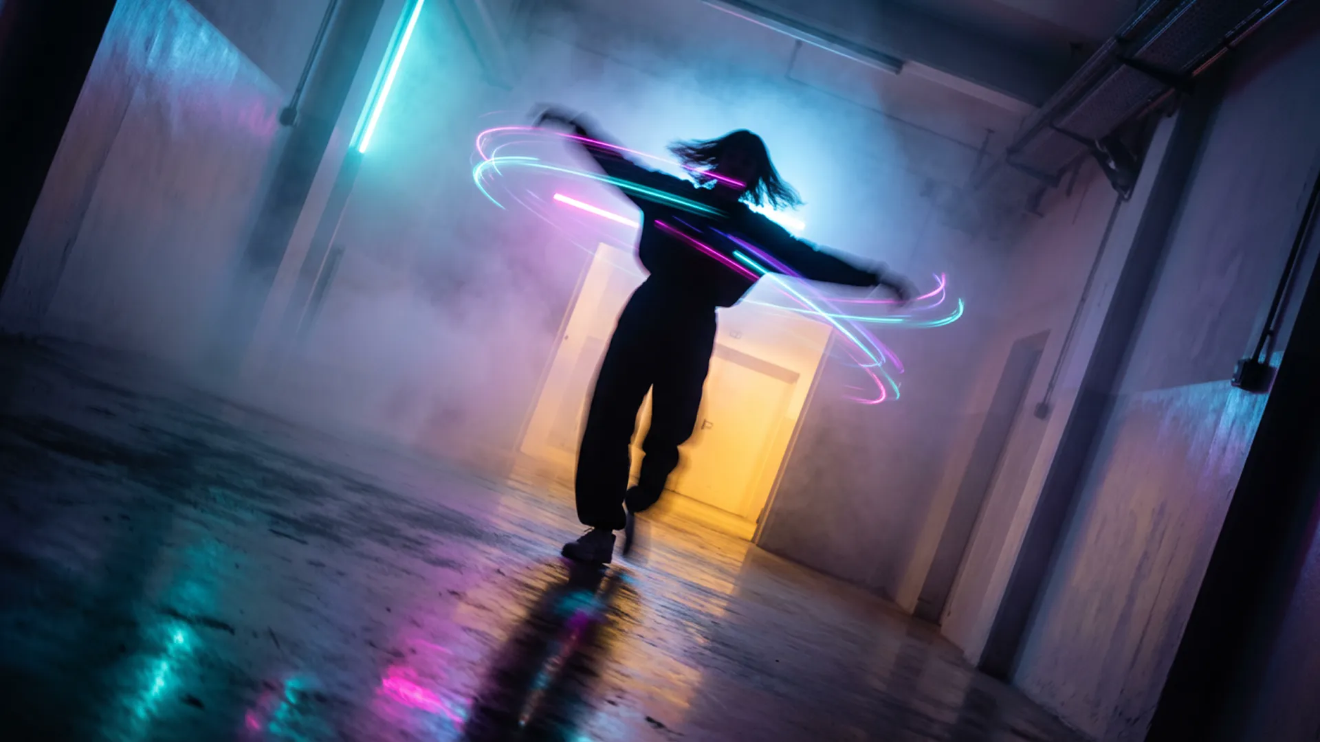 In a burst of uninhibited movement, a figure is captured mid-spin in a corridor of light and mist. The photography freezes the action at its peak, the person’s arms flung wide, their head tilted back in a moment of pure, kinetic release. This rapid twirl has visibly disturbed the dense, colored fog, creating eddies and vortices of cyan, magenta, and violet light around their silhouetted form. The composition is vibrant and chaotic, a stark contrast to a static scene. Light from a warm, yellow-orange portal ahead illuminates the swirling mist from one direction, while a cool blue glow from behind adds depth and complexity to the color palette. The floor, a dark mirror, reflects a blurred, chaotic version of the scene—a painterly smear of motion and color. The camera angle is slightly tilted, a Dutch angle that enhances the feeling of disequilibrium and exhilaration, capturing a fleeting, joyful rebellion against the quiet, contemplative nature of the space